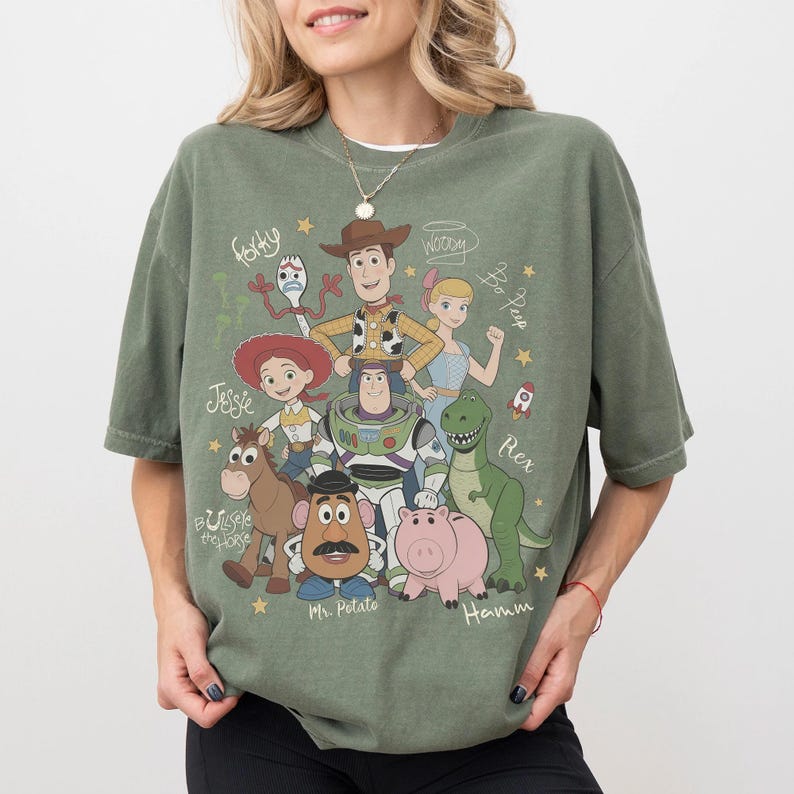 May include: An olive green short-sleeve t-shirt featuring a graphic print of Toy Story characters including Woody, Buzz Lightyear, Jessie, Bullseye, Mr. Potato Head, Hamm, Rex, Bo Peep, and Forky, with their names written in a cursive script font.