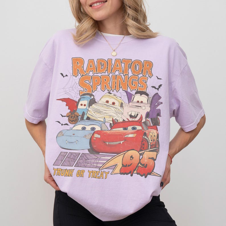 May include: A light purple t-shirt featuring a graphic of characters from the movie Cars, dressed in Halloween costumes. The text "Radiator Springs" and "Trunk or Treat" are displayed above and below the graphic, with the number "95" on the bottom right.