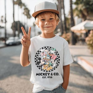 Puede incluir: Una camiseta blanca con un gr&aacute;fico circular de Mickey Mouse y sus amigos, sobre un fondo a cuadros blanco y negro. El texto "MICKEY & CO. EST. 1928" est&aacute; impreso debajo. El ni&ntilde;o lleva una gorra de b&eacute;isbol gris y marr&oacute;n.