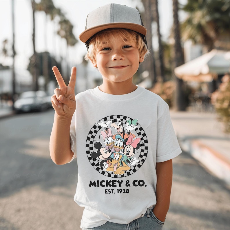 Puede incluir: Una camiseta blanca con un gr&aacute;fico circular de Mickey Mouse y sus amigos, sobre un fondo a cuadros blanco y negro. El texto "MICKEY & CO. EST. 1928" est&aacute; impreso debajo. El ni&ntilde;o lleva una gorra de b&eacute;isbol gris y marr&oacute;n.