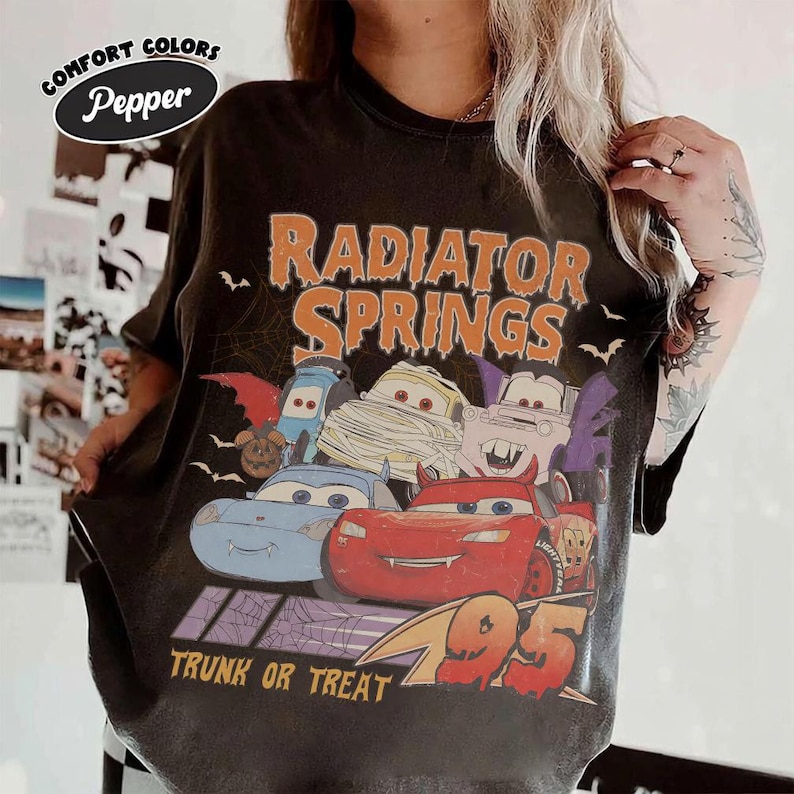 May include: Dark gray t-shirt featuring a Halloween-themed graphic of characters from the movie Cars, with the text "Radiator Springs" in orange. The design includes bats, a spiderweb, and the phrase "Trunk or Treat".