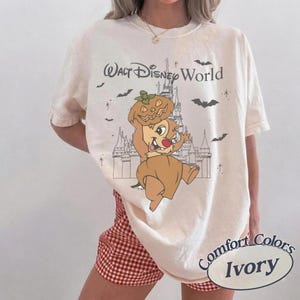 May include: Ivory-coloured t-shirt with a Halloween-themed graphic featuring a cartoon character wearing a pumpkin hat, with the text "Walt Disney World". The shirt is a Comfort Colors brand.