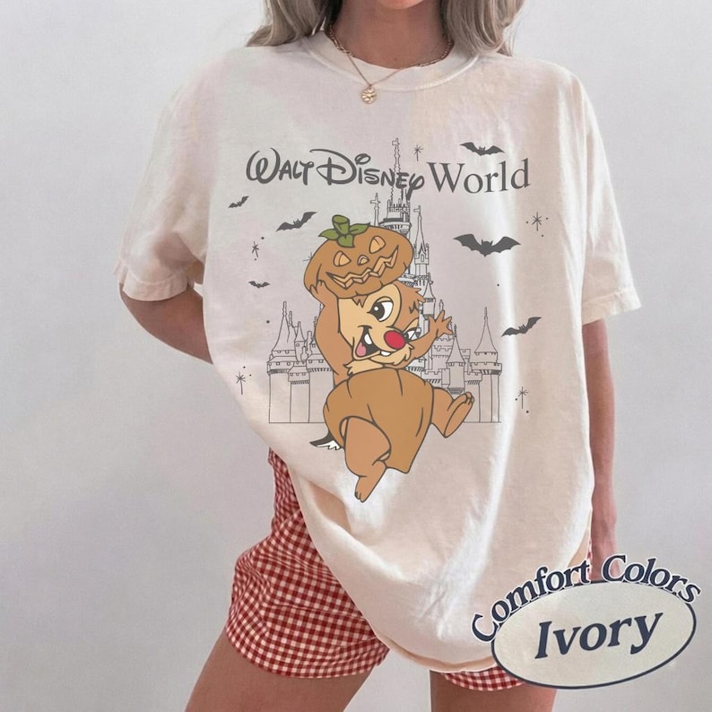 May include: Ivory-coloured t-shirt with a Halloween-themed graphic featuring a cartoon character wearing a pumpkin hat, with the text "Walt Disney World". The shirt is a Comfort Colors brand.