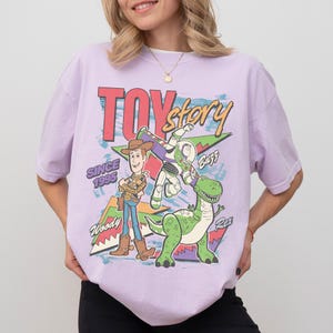 May include: A lilac t-shirt with a colourful graphic of Woody, Buzz Lightyear, and Rex from Toy Story. The words "Toy Story" and "Since 1995" are printed on the shirt in a retro style.