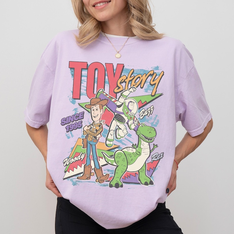May include: A lilac t-shirt with a colourful graphic of Woody, Buzz Lightyear, and Rex from Toy Story. The words "Toy Story" and "Since 1995" are printed on the shirt in a retro style.
