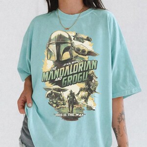 K&ouml;nnte beinhalten: Hellblaues T-Shirt mit einem Grafikdesign aus der Star Wars-Serie "The Mandalorian and Grogu". Das Design zeigt den Helm des Mandalorianers, Grogu und den Satz "THIS IS THE WAY".