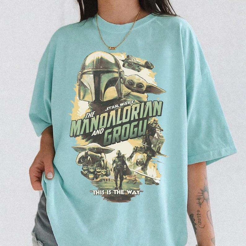 K&ouml;nnte beinhalten: Hellblaues T-Shirt mit einem Grafikdesign aus der Star Wars-Serie "The Mandalorian and Grogu". Das Design zeigt den Helm des Mandalorianers, Grogu und den Satz "THIS IS THE WAY".