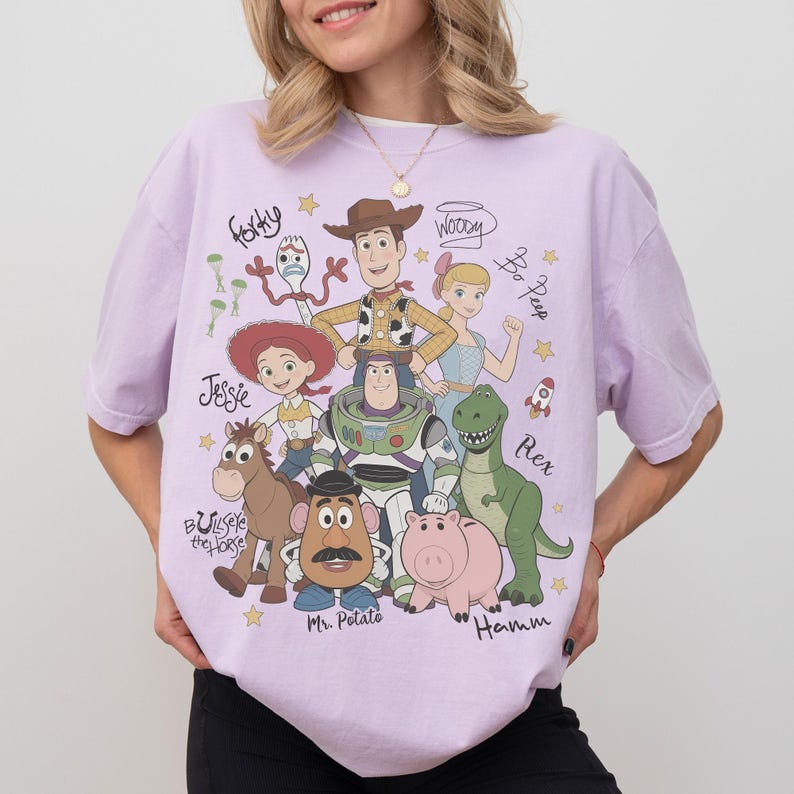 May include: A light purple t-shirt featuring a graphic print of Toy Story characters including Woody, Buzz Lightyear, Jessie, Bullseye, Mr. Potato Head, Hamm, Rex, Bo Peep, and Forky, with their names written in cursive script and small yellow stars.