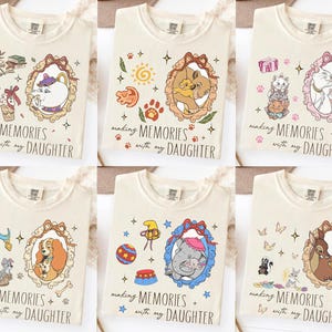 Op de afbeelding: Cr&egrave;mekleurige T-shirts met verschillende cartoonpersonages in sierlijke frames. Elk shirt heeft de tekst "making MEMORIES with my DAUGHTER". De ontwerpen tonen personages uit populaire animatiefilms.