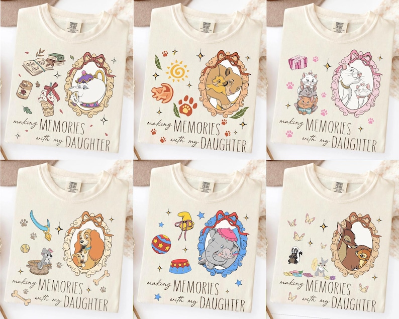 Op de afbeelding: Cr&egrave;mekleurige T-shirts met verschillende cartoonpersonages in sierlijke frames. Elk shirt heeft de tekst "making MEMORIES with my DAUGHTER". De ontwerpen tonen personages uit populaire animatiefilms.