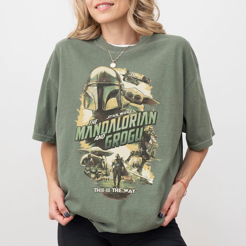 K&ouml;nnte beinhalten: Olivgr&uuml;nes T-Shirt mit einem Grafikdesign aus der Star Wars-Serie, "The Mandalorian and Grogu". Das Design zeigt den Helm des Mandalorianers, Grogu und den Satz "This is the Way".