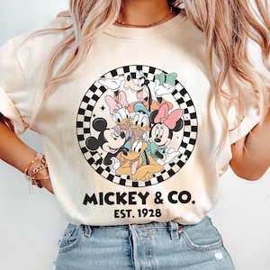 Puede incluir: Sudadera de color crema con un gr&aacute;fico circular de personajes de Disney, incluyendo Mickey Mouse, Minnie Mouse, Donald Duck y Goofy, rodeados por un borde a cuadros blanco y negro. El texto "MICKEY & CO. EST. 1928" est&aacute; impreso debajo.