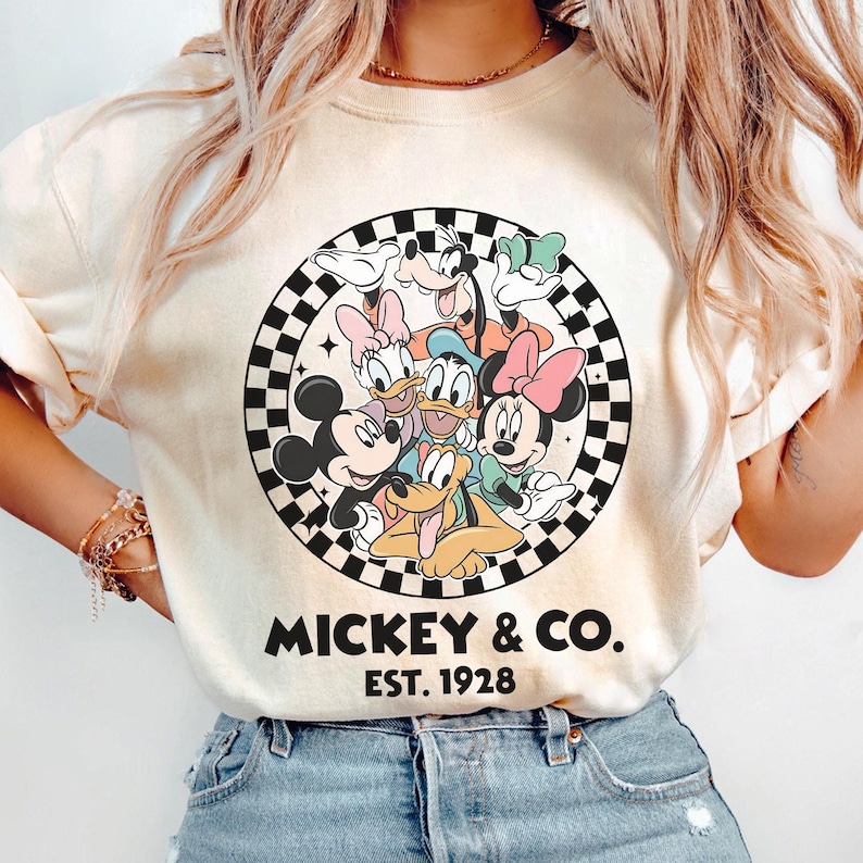 Puede incluir: Sudadera de color crema con un gr&aacute;fico circular de personajes de Disney, incluyendo Mickey Mouse, Minnie Mouse, Donald Duck y Goofy, rodeados por un borde a cuadros blanco y negro. El texto "MICKEY & CO. EST. 1928" est&aacute; impreso debajo.