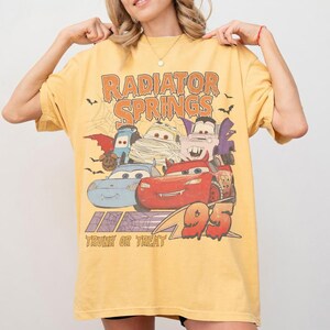 May include: Yellow t-shirt featuring a graphic of Cars characters in Halloween costumes with the text "Radiator Springs" and "Trunk or Treat 95". The design includes bats and a spiderweb.