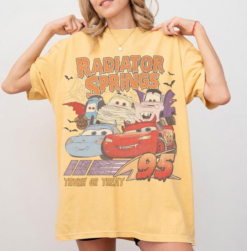 May include: Yellow t-shirt featuring a graphic of Cars characters in Halloween costumes with the text "Radiator Springs" and "Trunk or Treat 95". The design includes bats and a spiderweb.