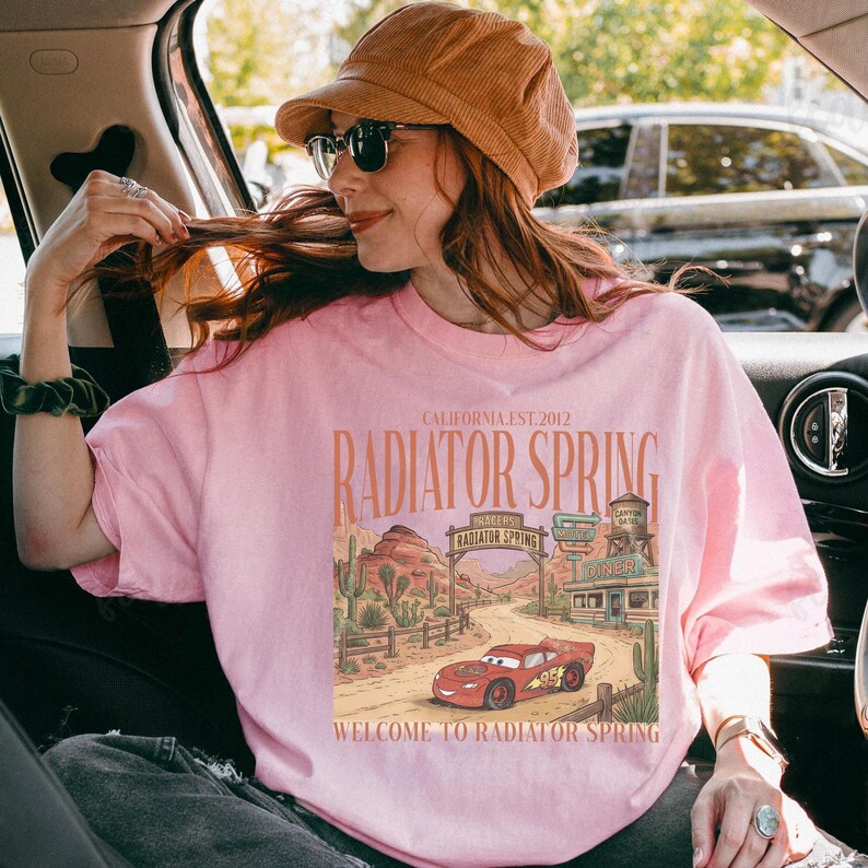 May include: A light pink t-shirt featuring a graphic of Radiator Springs, with the text "CALIFORNIA EST. 2012" and "RADIATOR SPRING". The graphic includes a red race car and desert scenery. The text "WELCOME TO RADIATOR SPRING" is also present.