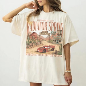 May include: Cream-colored t-shirt featuring a graphic of a red race car in front of a desert landscape with the text "Radiator Spring" and "Welcome to Radiator Spring". The shirt has short sleeves and a relaxed fit.