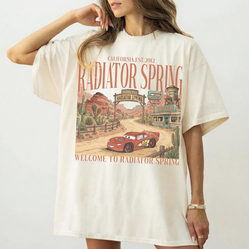 May include: Cream-colored t-shirt featuring a graphic of a red race car in front of a desert landscape with the text "Radiator Spring" and "Welcome to Radiator Spring". The shirt has short sleeves and a relaxed fit.
