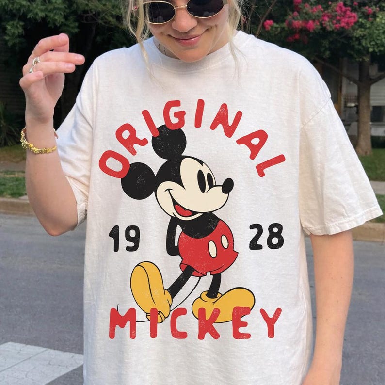 May include: Cream-colored t-shirt featuring a classic Mickey Mouse graphic. The design includes the word "ORIGINAL" in red, the year "1928", and the name "MICKEY" in red. The cartoon character is depicted in his iconic red shorts and yellow shoes.