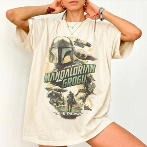 K&ouml;nnte beinhalten: Cremefarbenes T-Shirt mit einer Grafik aus der Serie "The Mandalorian". Das Design zeigt den Mandalorianer-Helm, Grogu und den Text "The Mandalorian and Grogu" und "This is the Way".