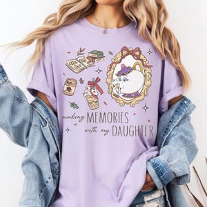 Op de afbeelding: Lichtpaars T-shirt met een grafisch ontwerp met een theepot, boeken en de tekst "making MEMORIES with my DAUGHTER". Het ontwerp bevat een ingelijste theepot en andere decoratieve elementen.