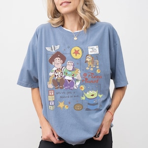 Pu&ograve; includere: T-shirt azzurra con una grafica colorata dei personaggi di Toy Story, tra cui Woody e Buzz Lightyear. Il design include il logo Pizza Planet e la frase "you've got a friend in me".