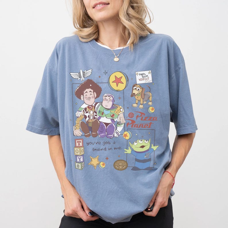 Pu&ograve; includere: T-shirt azzurra con una grafica colorata dei personaggi di Toy Story, tra cui Woody e Buzz Lightyear. Il design include il logo Pizza Planet e la frase "you've got a friend in me".