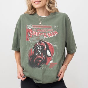 Może przedstawiać: Oliwkowo-zielona koszulka z nadrukiem w stylu vintage z okładki komiksu Spider-Man. Grafika zawiera tytuł "The Amazing Spider-Man" w kolorze czerwono-białym, ze szczeg&oacute;łową ilustracją twarzy superbohatera.