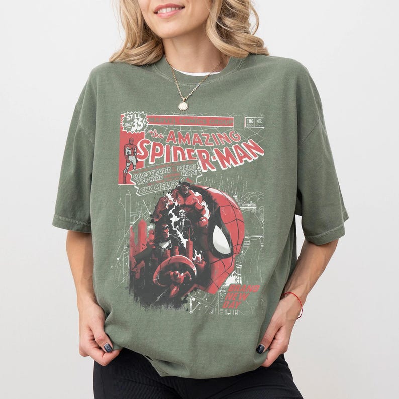 Może przedstawiać: Oliwkowo-zielona koszulka z nadrukiem w stylu vintage z okładki komiksu Spider-Man. Grafika zawiera tytuł "The Amazing Spider-Man" w kolorze czerwono-białym, ze szczeg&oacute;łową ilustracją twarzy superbohatera.