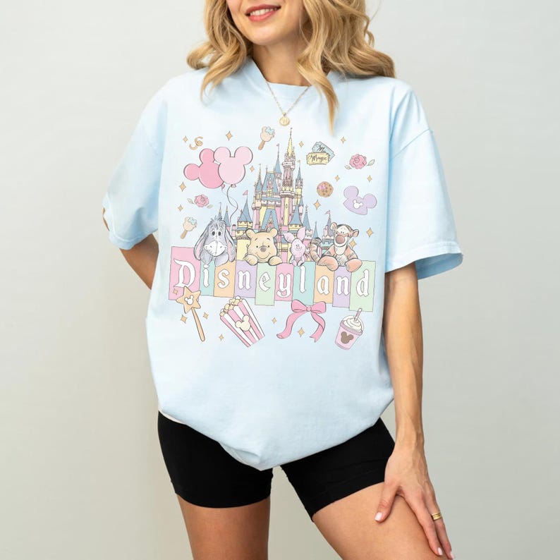 Puede incluir: Camiseta azul claro con un gr&aacute;fico de Disneyland. El dise&ntilde;o incluye un castillo, personajes de dibujos animados y la palabra "Disneyland" en colores pastel. Gr&aacute;ficos adicionales incluyen un globo rosa, palomitas de ma&iacute;z y un lazo.