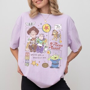 Pu&ograve; includere: T-shirt lilla chiaro con una grafica colorata dei personaggi di Toy Story, tra cui Woody, Buzz Lightyear, Slinky Dog e i piccoli omini verdi. Il design include anche il testo "you've got a friend in me" e "Pizza Planet".