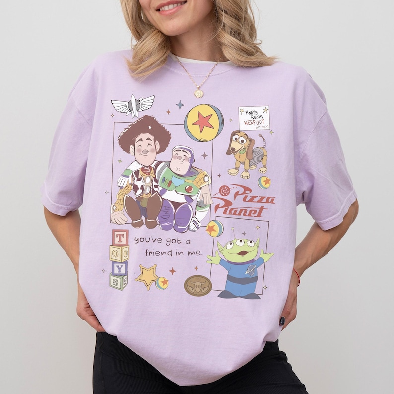 Pu&ograve; includere: T-shirt lilla chiaro con una grafica colorata dei personaggi di Toy Story, tra cui Woody, Buzz Lightyear, Slinky Dog e i piccoli omini verdi. Il design include anche il testo "you've got a friend in me" e "Pizza Planet".