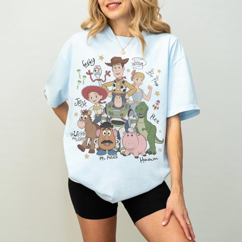 May include: A light blue t-shirt featuring a graphic print of Toy Story characters including Woody, Buzz Lightyear, Jessie, Bullseye, Mr. Potato Head, Hamm, Rex, Bo Peep, and Forky, with their names written in a cursive script alongside small gold stars.