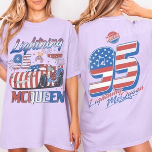 Może przedstawiać: Jasnofioletowy t-shirt z grafiką Lightning McQueen z przodu oraz napisem Lightning 95 McQueen. Z tyłu znajduje się duża liczba 95 w czerwono-biało-niebieski wz&oacute;r flagi z napisem Lightning McQueen oraz logo Rust-eze.