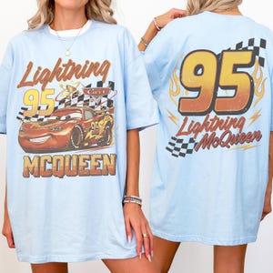 May include: Pale blue t-shirt with a Lightning McQueen graphic. The front shows a race car, the number 95, and the text "Lightning McQueen". The back has "95 Lightning McQueen" with flames and a checkered flag design.