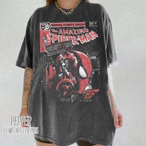 Może przedstawiać: Ciemnoszara koszulka z okładką komiksu The Amazing Spider-Man. Projekt zawiera twarz Spider-Mana, kolory czerwony i czarny oraz tekst komiksowy. Koszulka ma kr&oacute;tkie rękawy i napis "PEPPER COMFORT COLORS".
