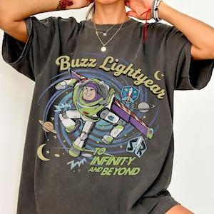 May include: A charcoal gray short-sleeve t-shirt featuring a graphic of Buzz Lightyear flying through space. The design includes the text Buzz Lightyear and To Infinity and Beyond, along with planets, stars, and a crescent moon in a colorful print.