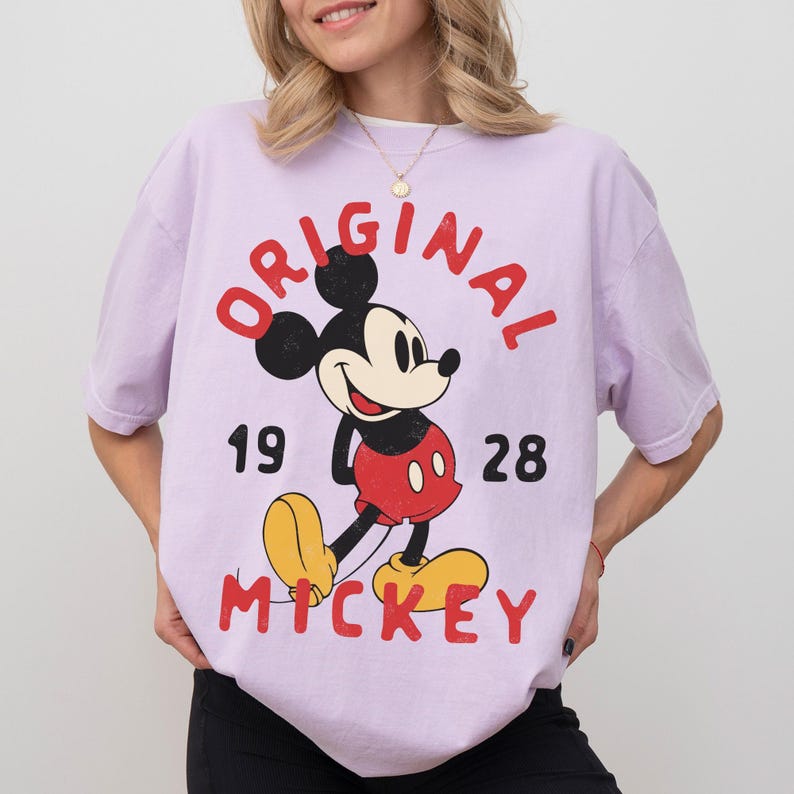 May include: A light purple t-shirt featuring a classic Mickey Mouse graphic. The design includes the word "ORIGINAL" in red, the year "1928", and the name "MICKEY". Mickey Mouse is depicted in his iconic red shorts, yellow shoes, and black gloves.