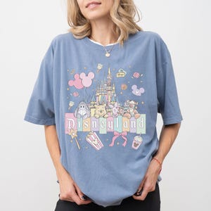 Puede incluir: Una camiseta azul claro con un dise&ntilde;o de Disneyland. El dise&ntilde;o incluye un castillo, personajes de dibujos animados y la palabra "Disneyland" en una fuente pastel. Otros elementos incluyen palomitas de ma&iacute;z, una varita m&aacute;gica y una bebida.