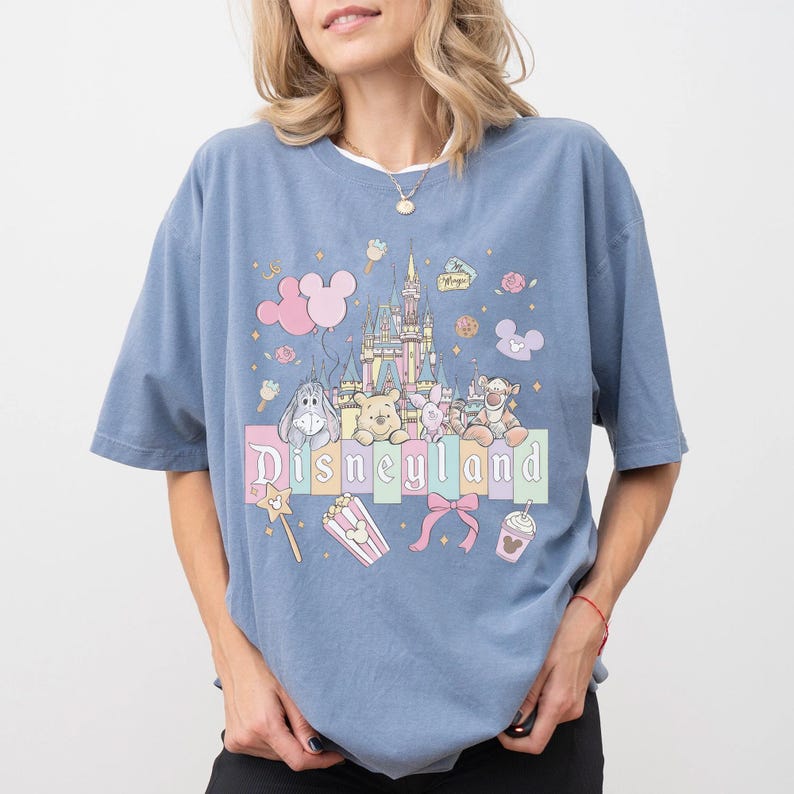 Puede incluir: Una camiseta azul claro con un dise&ntilde;o de Disneyland. El dise&ntilde;o incluye un castillo, personajes de dibujos animados y la palabra "Disneyland" en una fuente pastel. Otros elementos incluyen palomitas de ma&iacute;z, una varita m&aacute;gica y una bebida.