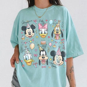 Puede incluir: Una camiseta azul claro con los personajes de dibujos animados Mickey Mouse, Daisy Duck, Pluto, Donald Duck, Goofy y Minnie Mouse. El dise&ntilde;o incluye gorros de fiesta, globos y el texto "Mickey Mouse" y "Daisy Duck".