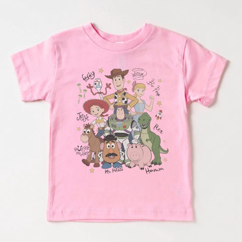 May include: A light pink short-sleeve t-shirt featuring a graphic print of Toy Story characters including Woody, Buzz Lightyear, Jessie, Bullseye, Mr. Potato Head, Rex, Hamm, Bo Peep, and Forky, with their names written in a cursive script font.