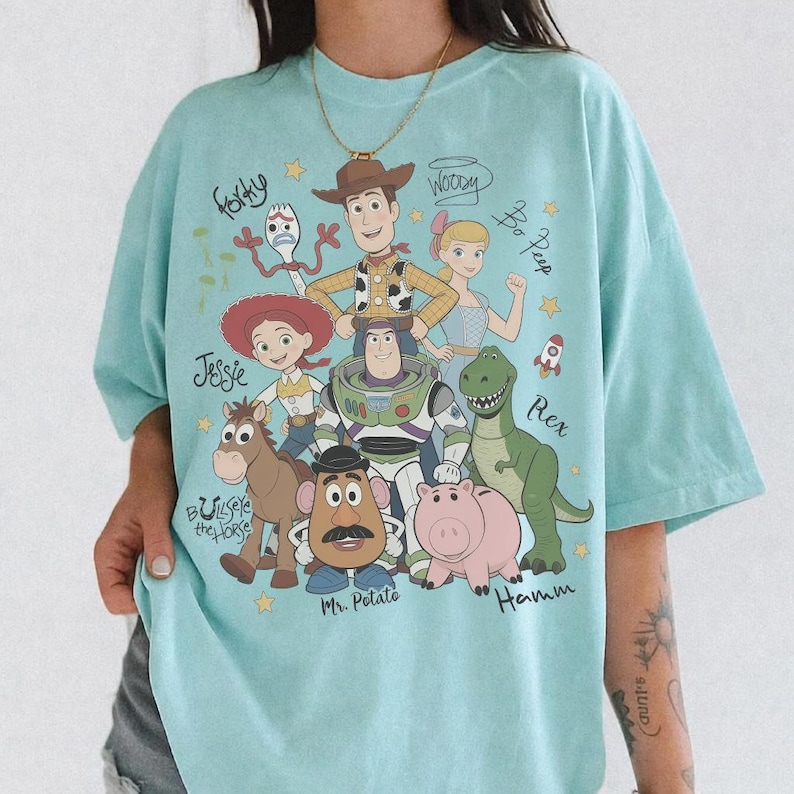 May include: A light blue t-shirt featuring a graphic print of Toy Story characters including Woody, Buzz Lightyear, Jessie, Bullseye, Mr. Potato Head, Rex, Hamm, Bo Peep, and Forky, with their names written in cursive script and small yellow stars.