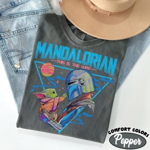 May include: A grey t-shirt with a graphic of The Mandalorian and Grogu, with the text "MANDALORIAN" in blue and "THIS IS THE WAY" in white. The shirt is styled with a tan hat and blue jeans.