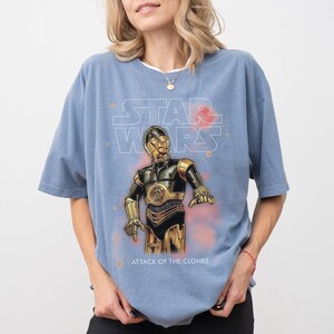 Peut inclure: Un t-shirt bleu clair avec un graphisme de C-3PO de Star Wars. Le texte "STAR WARS" est au-dessus du personnage, et "ATTACK OF THE CLONES" est en dessous. Le design comprend des accents rouges et oranges.