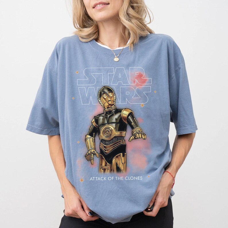 Peut inclure: Un t-shirt bleu clair avec un graphisme de C-3PO de Star Wars. Le texte "STAR WARS" est au-dessus du personnage, et "ATTACK OF THE CLONES" est en dessous. Le design comprend des accents rouges et oranges.