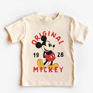 May include: Cream-colored t-shirt featuring a vintage Mickey Mouse graphic. The design includes the words "ORIGINAL MICKEY" in red, with the numbers "1928" below. Mickey Mouse is depicted in his classic red shorts, yellow shoes, and black ears.