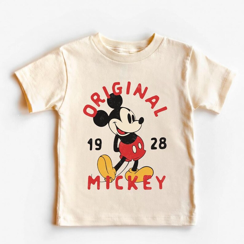 May include: Cream-colored t-shirt featuring a vintage Mickey Mouse graphic. The design includes the words "ORIGINAL MICKEY" in red, with the numbers "1928" below. Mickey Mouse is depicted in his classic red shorts, yellow shoes, and black ears.