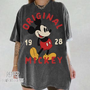 May include: Dark gray t-shirt featuring a vintage Mickey Mouse graphic. The design includes the words "ORIGINAL MICKEY" in red, with the year "1928". The shirt is from the brand "PEPPER COMFORT COLORS".