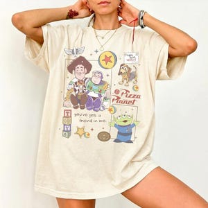 Pu&ograve; includere: T-shirt color crema con personaggi dei cartoni animati del film Toy Story. Il design include Woody, Buzz Lightyear, un cane e un alieno, con la scritta "Pizza Planet" e "You've got a friend in me."